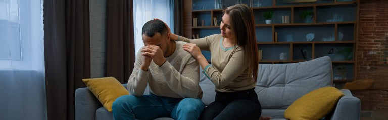 As they are seated in their home, a wife places her hands on her husband's shoulder while he is struggling to forgive her.